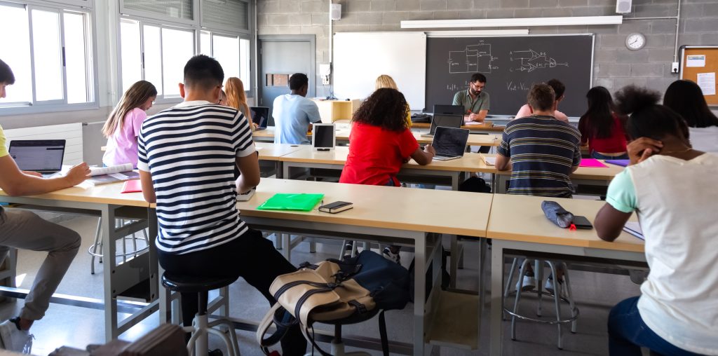 Rear view of group of multiracial high school students in class using laptops and doing homework while teacher marks exams in red pen. Education.