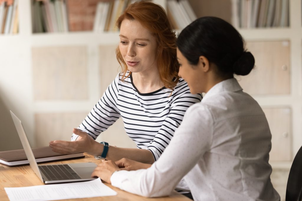 Manager provides professional aid, consult Indian female client use laptop show online presentation, convince buy services of company during meeting in office. Mentor helps new worker with app concept