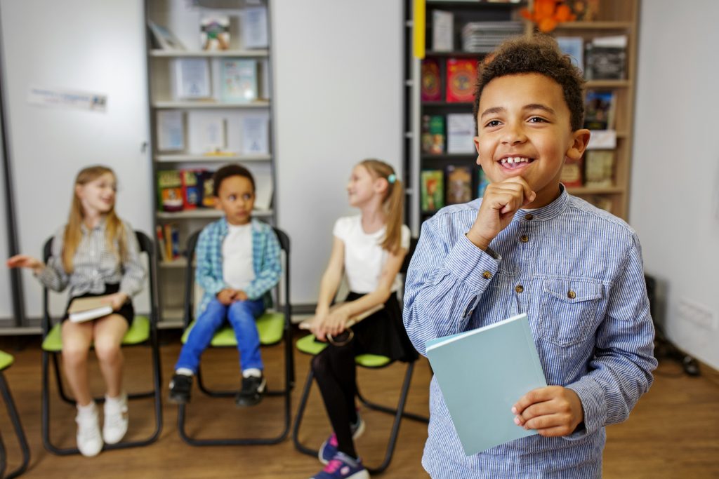 Afro american schoolboy standing in middle of classroom and answering question in front of classmates and teacher.