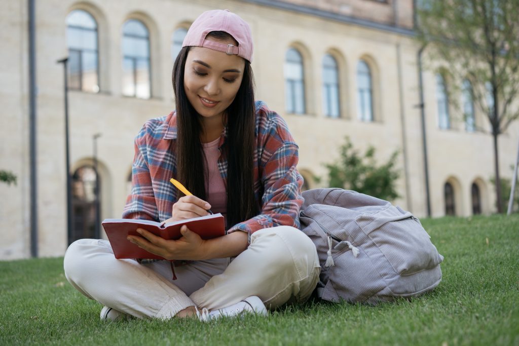 Smiling asian student studying, exam preparation, sitting on grass in university campus. Young woman taking notes working outdoors. Education concept