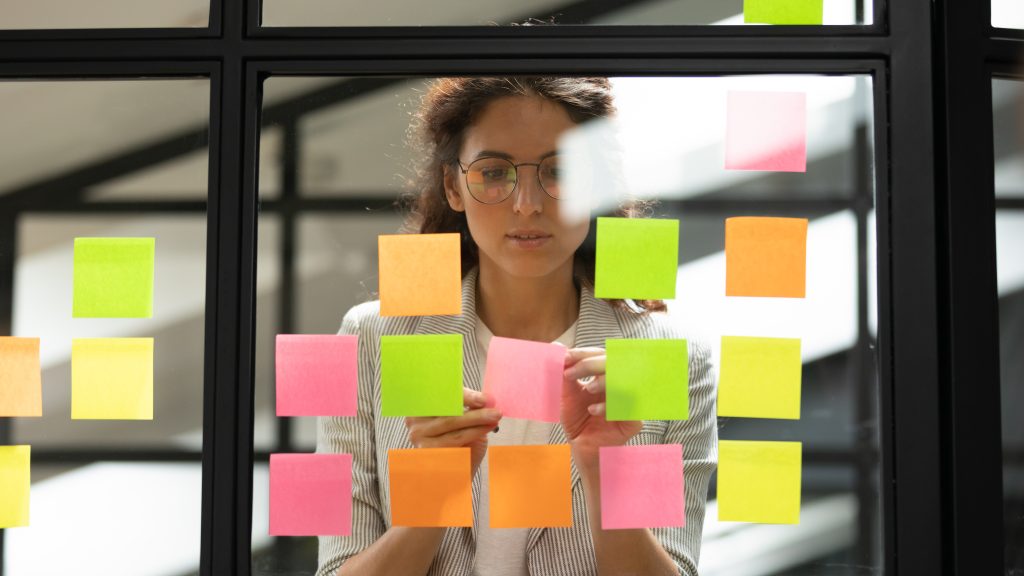 Attractive concentrated business lady in glasses creating to-do list using multi coloured post-it sticky notes attaching them to transparent wall standing behind glass view, be more productive concept