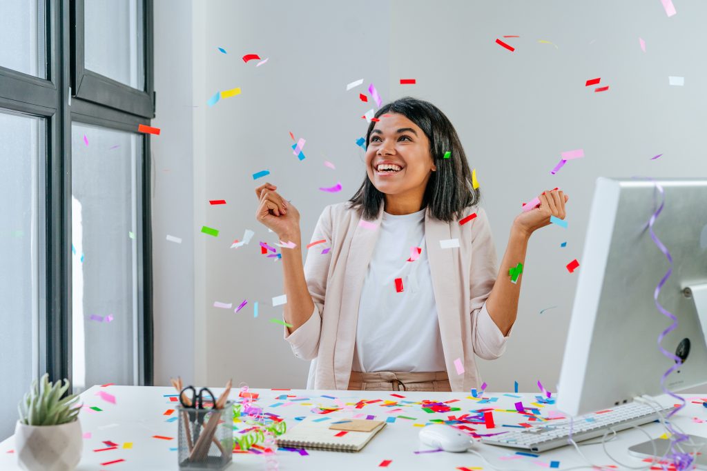 Young business woman having fun time catching confetti sitting at the desk in the office. Party time on the work place. Selective focus.