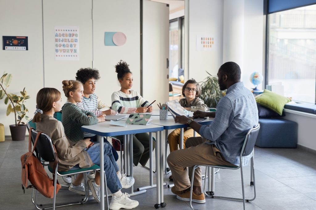 Full length shot of diverse group of children sitting at table with male teacher in modern school classroom