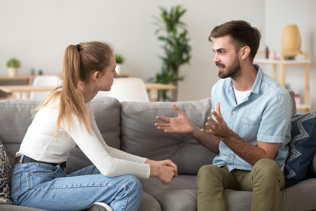 Serious young couple sitting together on sofa, talking about relationships, spending time together at home, focused wife listening to speaking husband, friends having conversation