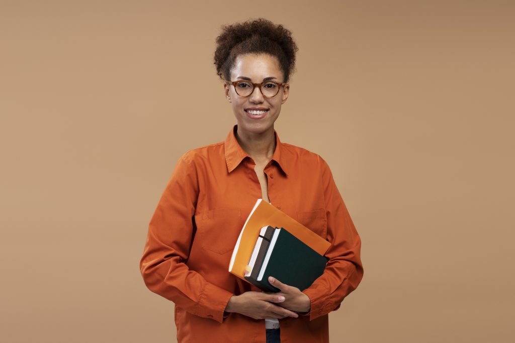 Confident young African American woman, high school teacher, tutor wearing glasses and orange casual shirt, posing with books, isolated on beige color background. College, Knowledge, Education concept