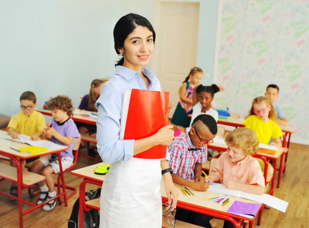 a young attractive female primary school teacher smiles at the background of school children sitting at their desks. Back to school, teacher's Day.