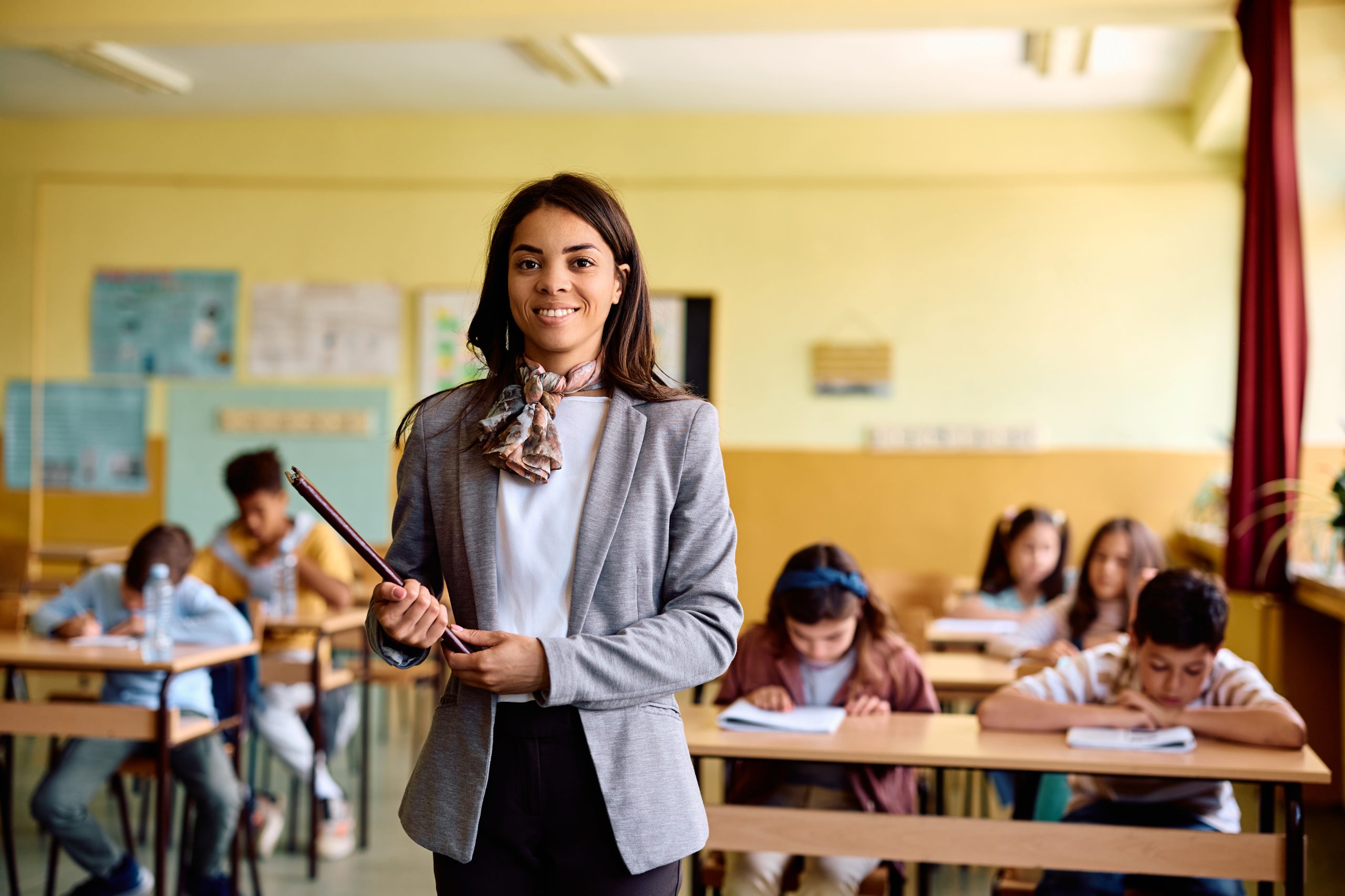 Happy Hispanic teacher during a class at elementary school looking at camera. her students are in the background.