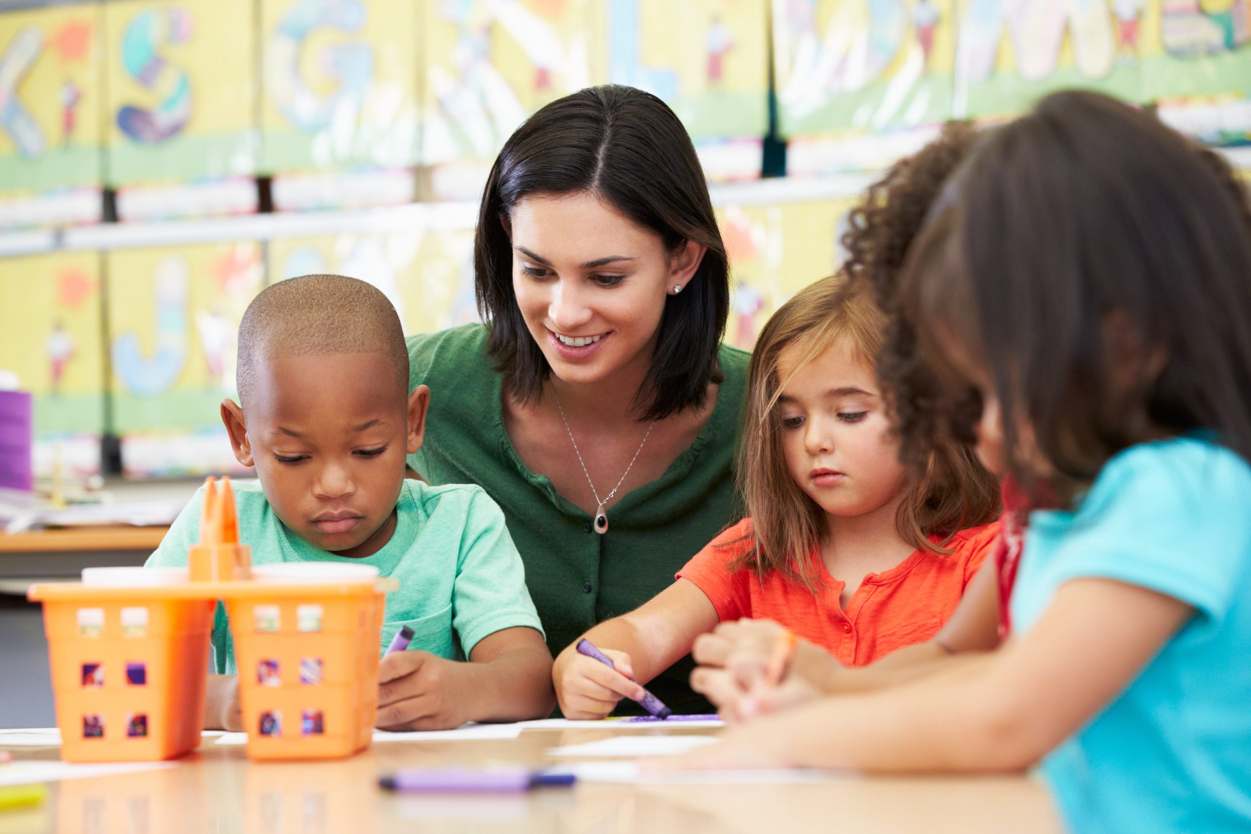 Group Of Elementary Age Children In Art Class With Teacher Sitting Down At Table Drawing