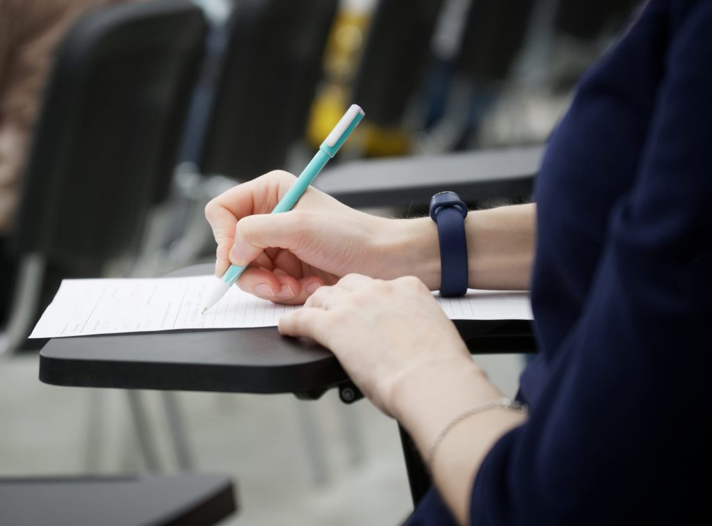 A girl writes a dictation or fills out documents in the audience, sitting on a school chair with a writing stand. Close-up. No face