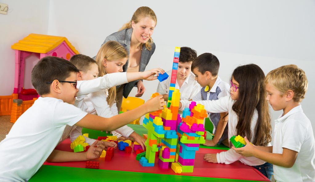 Happy kids and female teacher playing together with colorful toy building blocks in classroom at elementary school