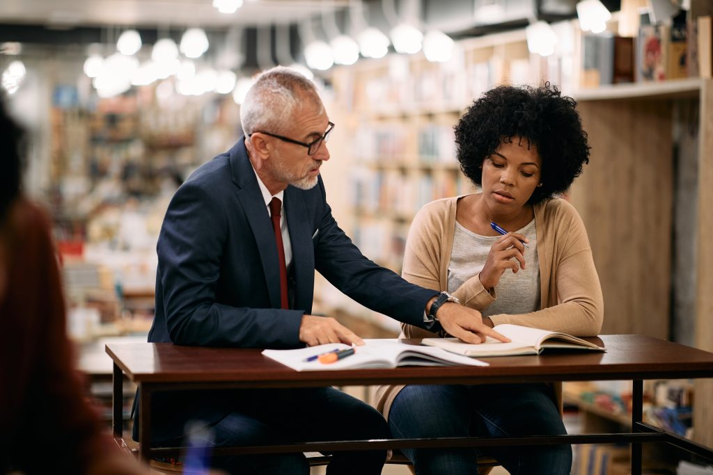 Mature professor helping to African American mid adult student while she is learning in a library.