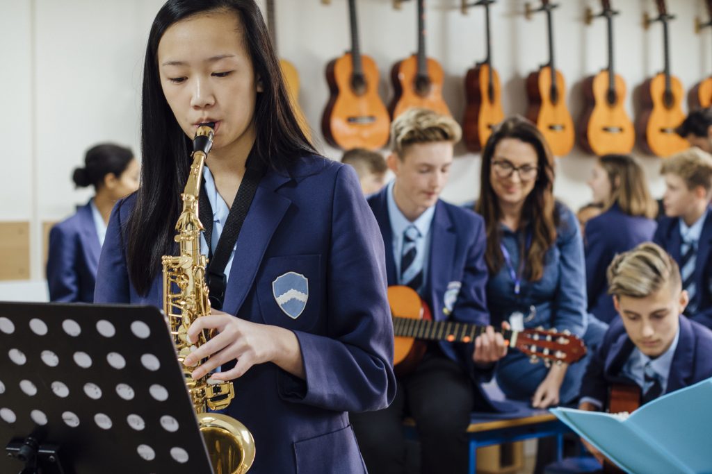 Teen student is playing the saxophone in her school music lesson. The rest of the class are in the background, out of focus.