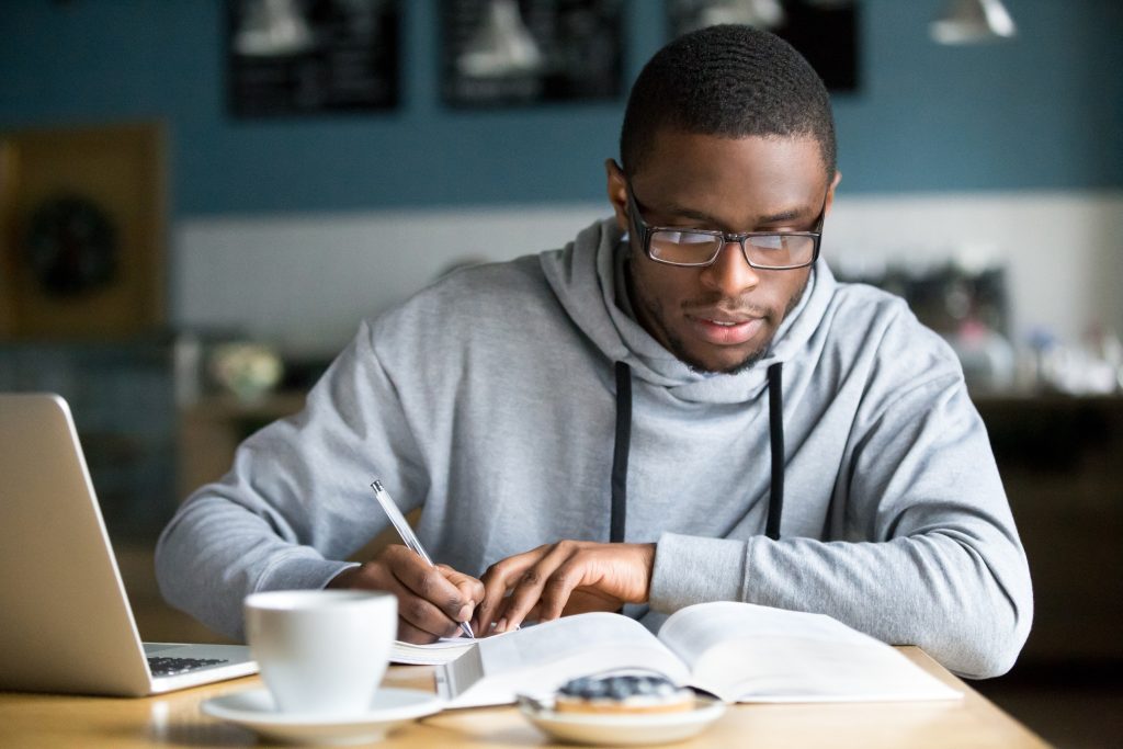 Focused millennial african american student in glasses making notes writing down information from book in cafe preparing for test or exam, young serious black man studying or working in coffee house