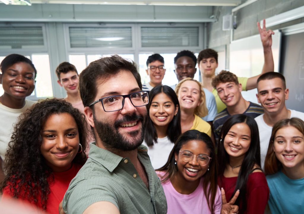 Happy selfie of young group of students taking a photo with their male teacher in the classroom, celebrating the end of course. Classmates from different countries, looking at camera with big smiles.