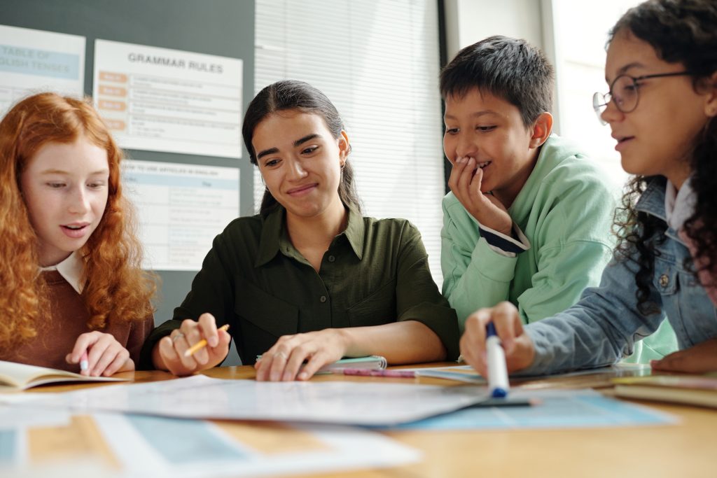 Group of clever schoolkids and their teacher discussing new grammar rules while looking at paper with English tenses or parts of speech at lesson