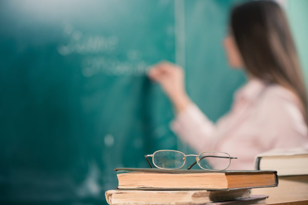 glasses and books at the classroom table while teacher writing on a blackboard