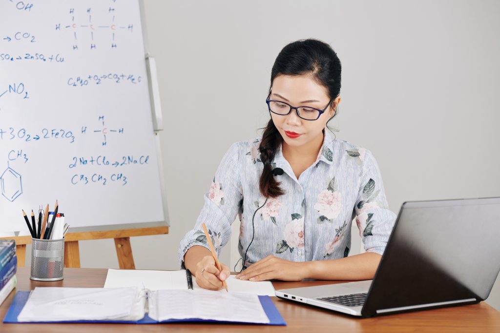 Young female Asian science teacher in glasses filling documents and making plan on online lesson