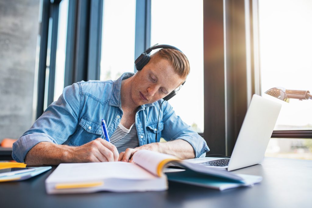 Shot of young man studying in college library sitting at table with laptop and books. Male university student preparing notes for the exam at library.