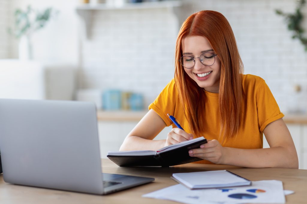 Happy young caucasian woman student using laptop computer for online education, watching the webinar, taking notes in a notebook, studying, distance learning at home