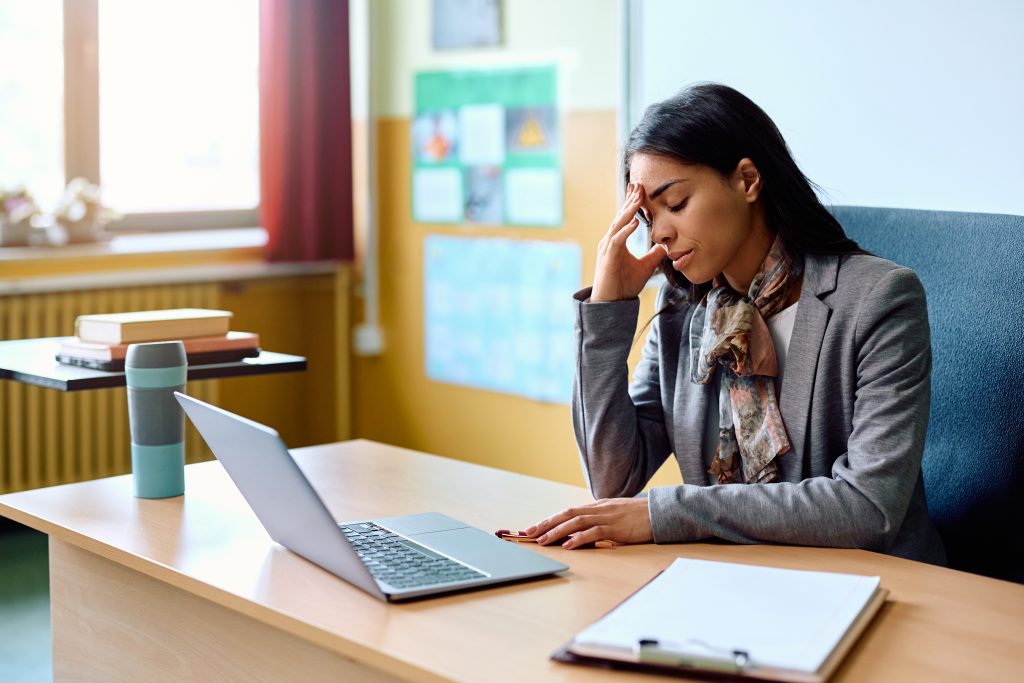 Hispanic teacher holding her head in pain while sitting at her desk in the classroom.