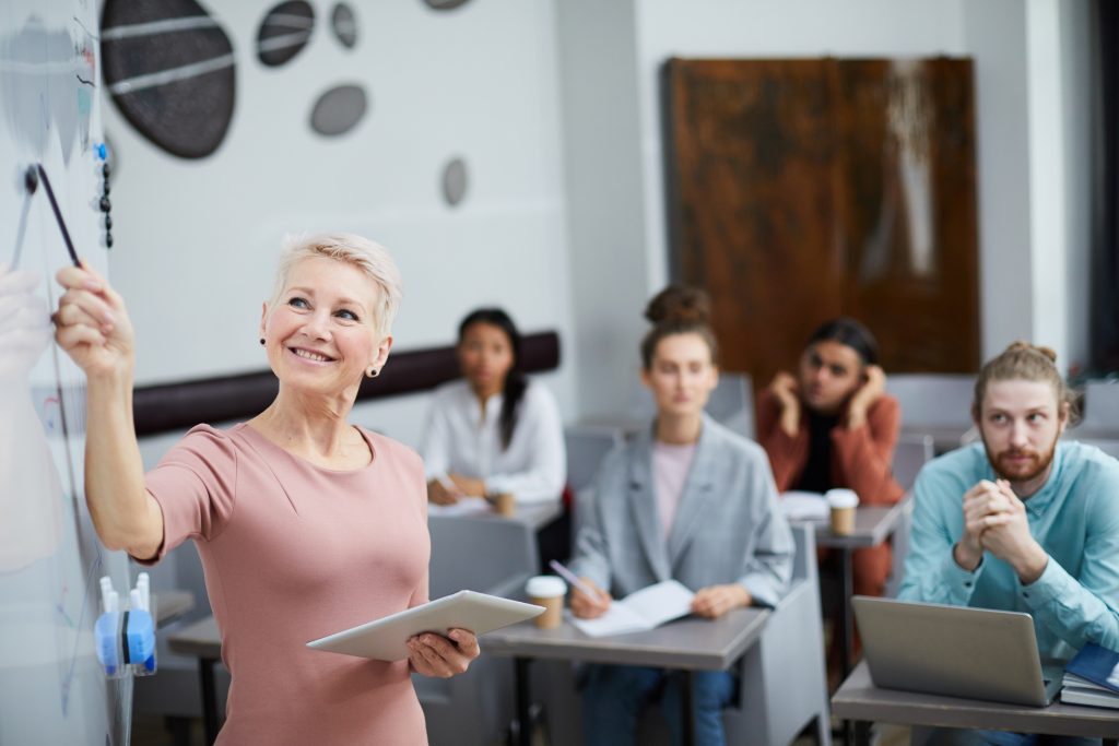 Waist up portrait of smiling female teacher pointing at whiteboard while presenting training course for students in class, copy space