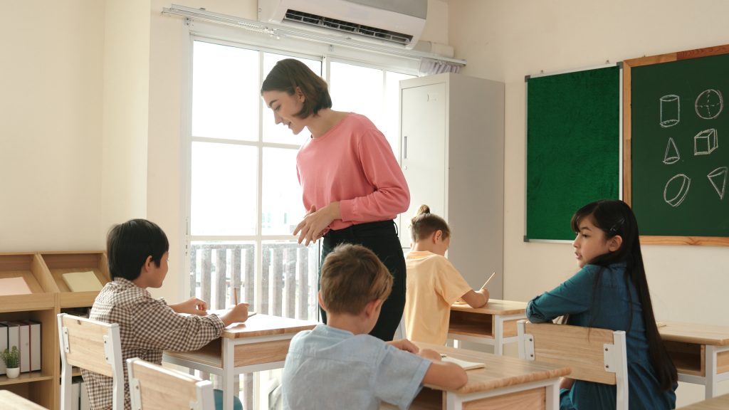 Caucasian teacher walking and check student homework at classroom while diverse children doing classwork at classroom. Diverse smart student lecturing and taking a note at elementary school. Pedagogy.