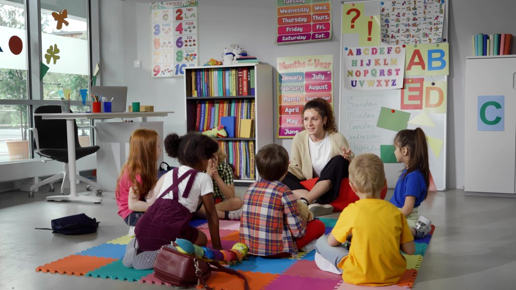 Small nursery school children with teacher sitting on floor having lesson. Young woman teach preschool kids in classroom sitting on floor together