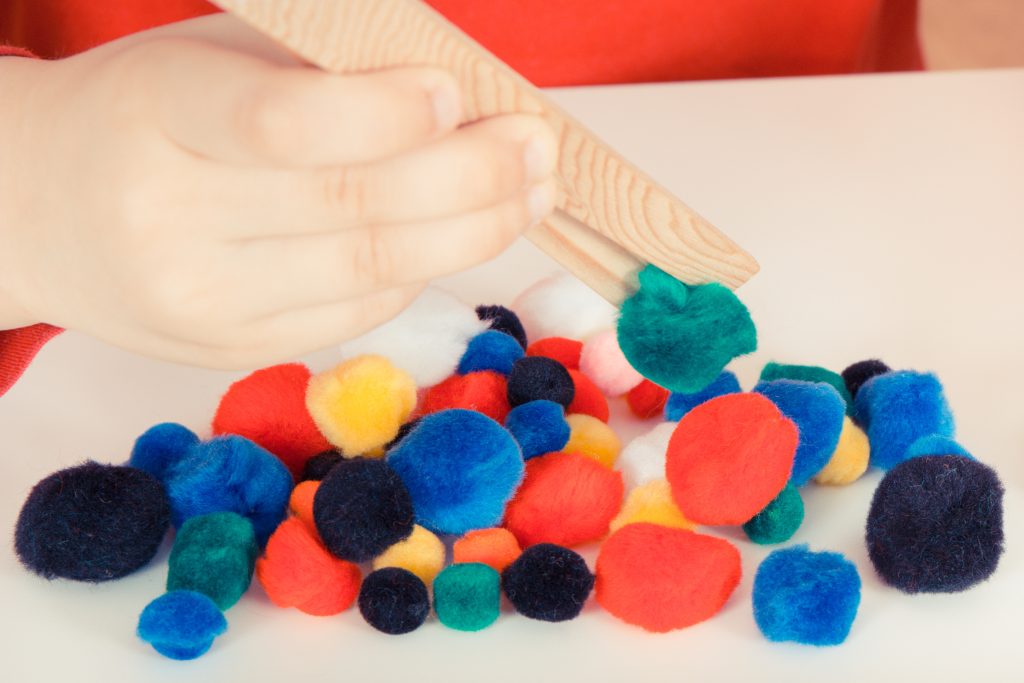 Preschooler playing with small fluffy colorful pompoms and wooden tongs. Development of kids motor skills, coordination, creativity and logical thinking