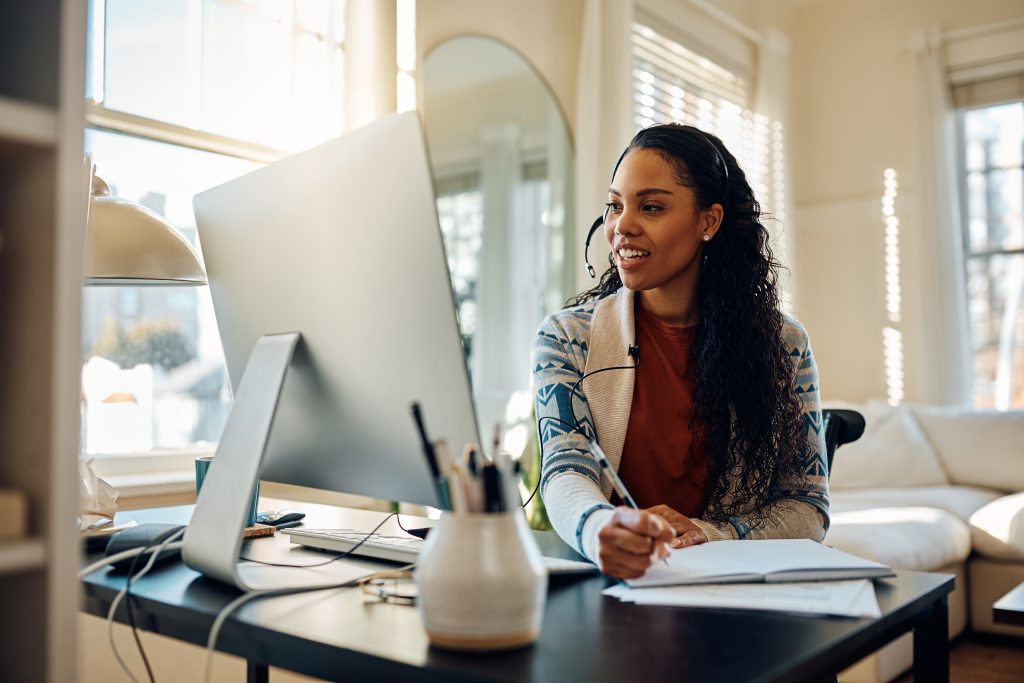 Happy African American woman using computer and taking notes while following online class from home.