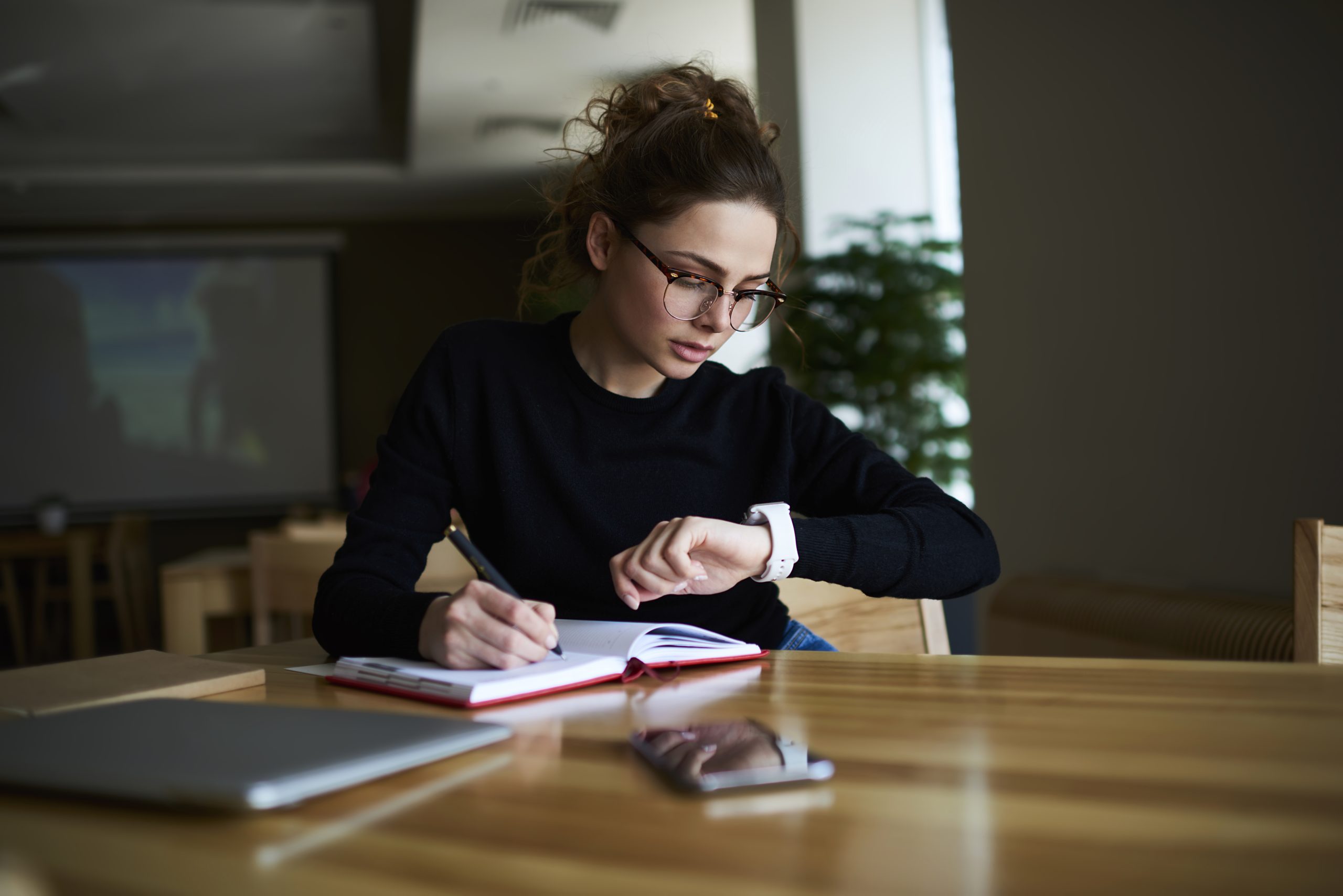 Talented female writer in stylish eyeglasses noting some information in notepad while looking on alarm watch to managing time for organization of working process sitting in university indoors