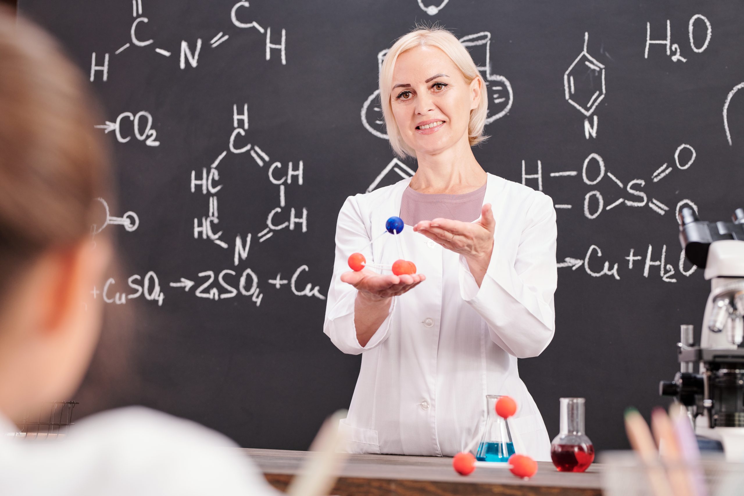 Smiling teacher of chemistry presenting model of molecule of chemical compound to schoolkids against blackboard with chemical formula