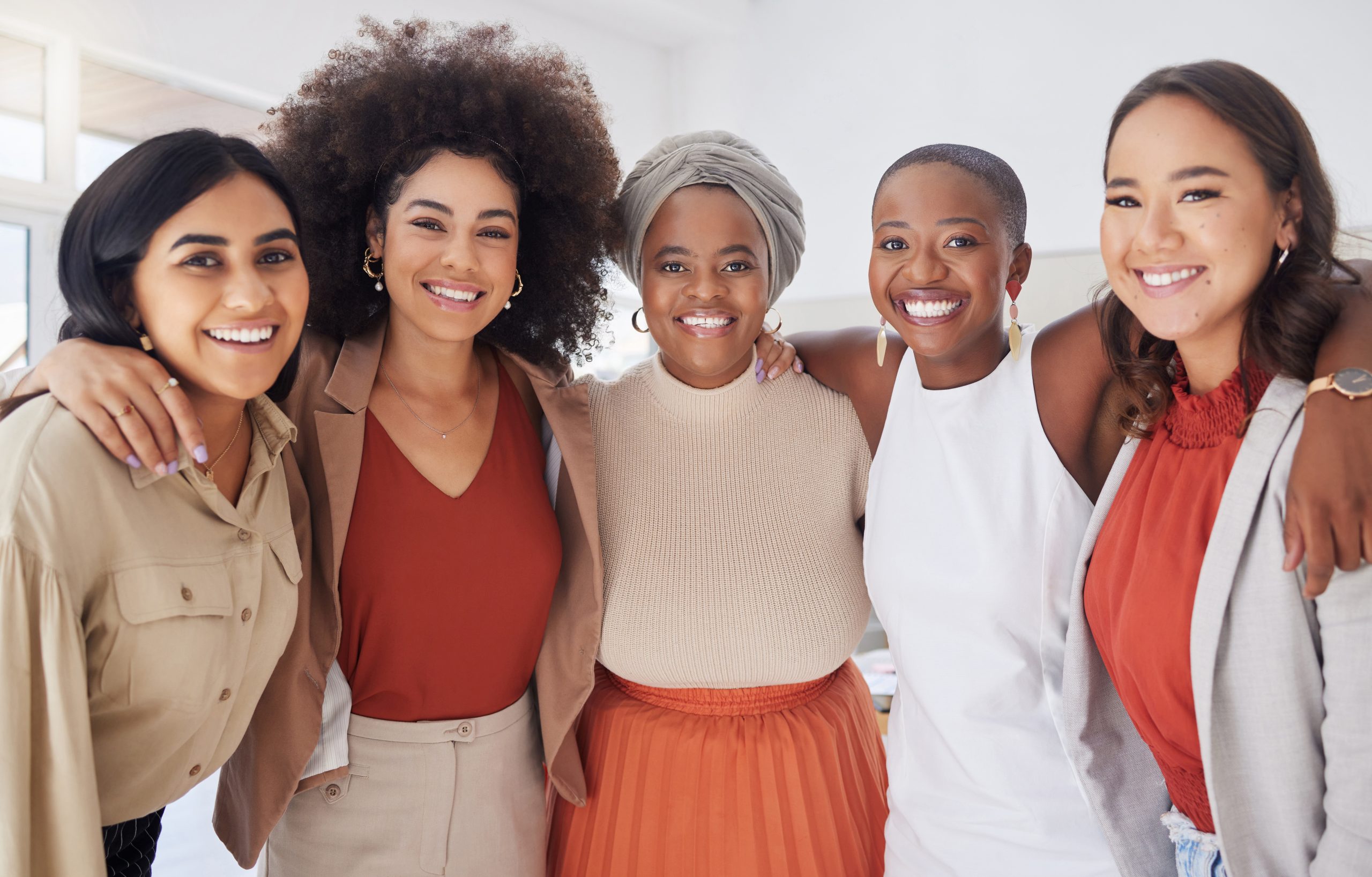 Portrait of a diverse group of smiling ethnic business women standing together in the office. Ambitious happy confident professional team of colleagues embracing while feeling supported and empowered