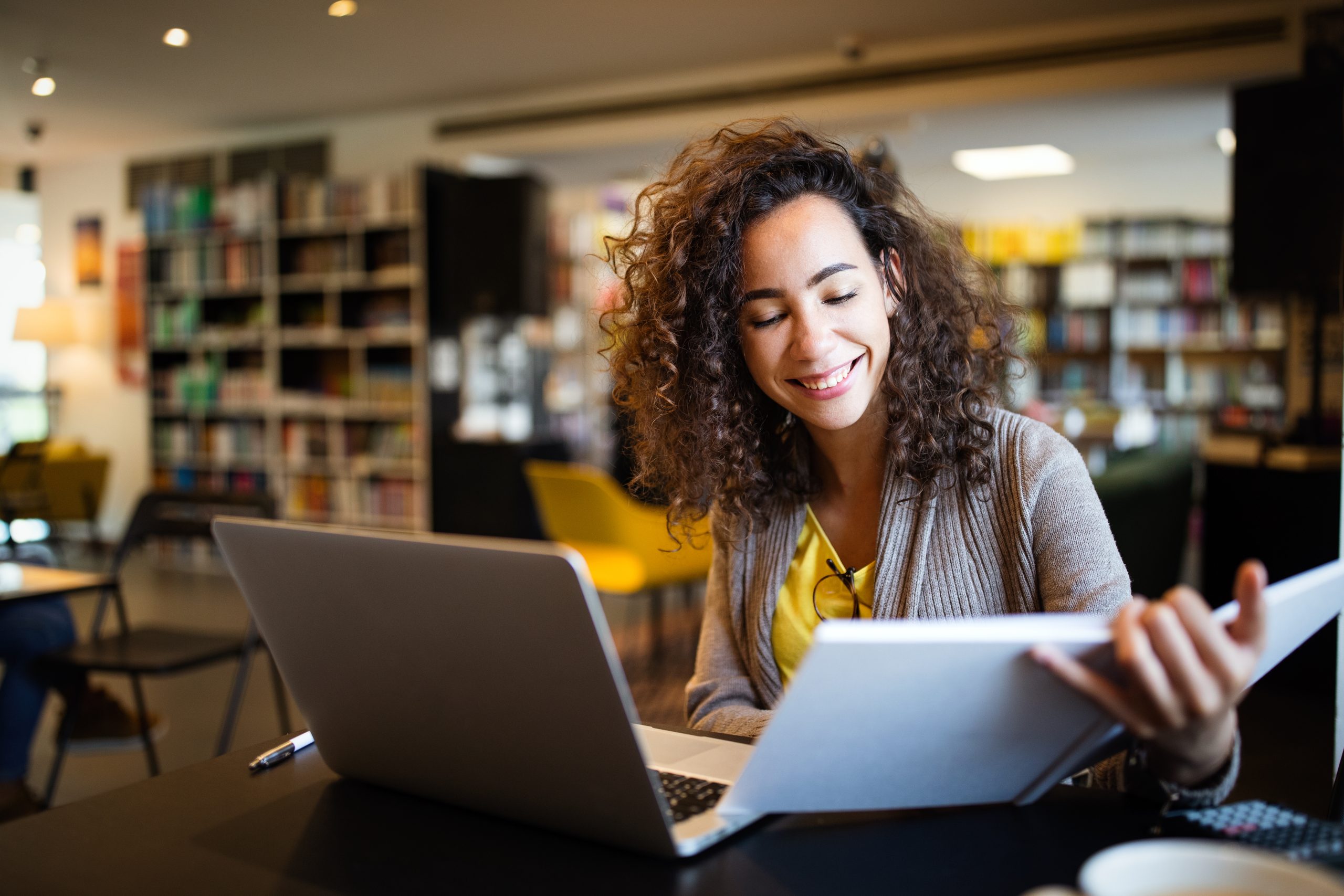 Young afro american woman sitting at table with books and laptop for finding information. Young student taking notes from laptop and books for her study in library.