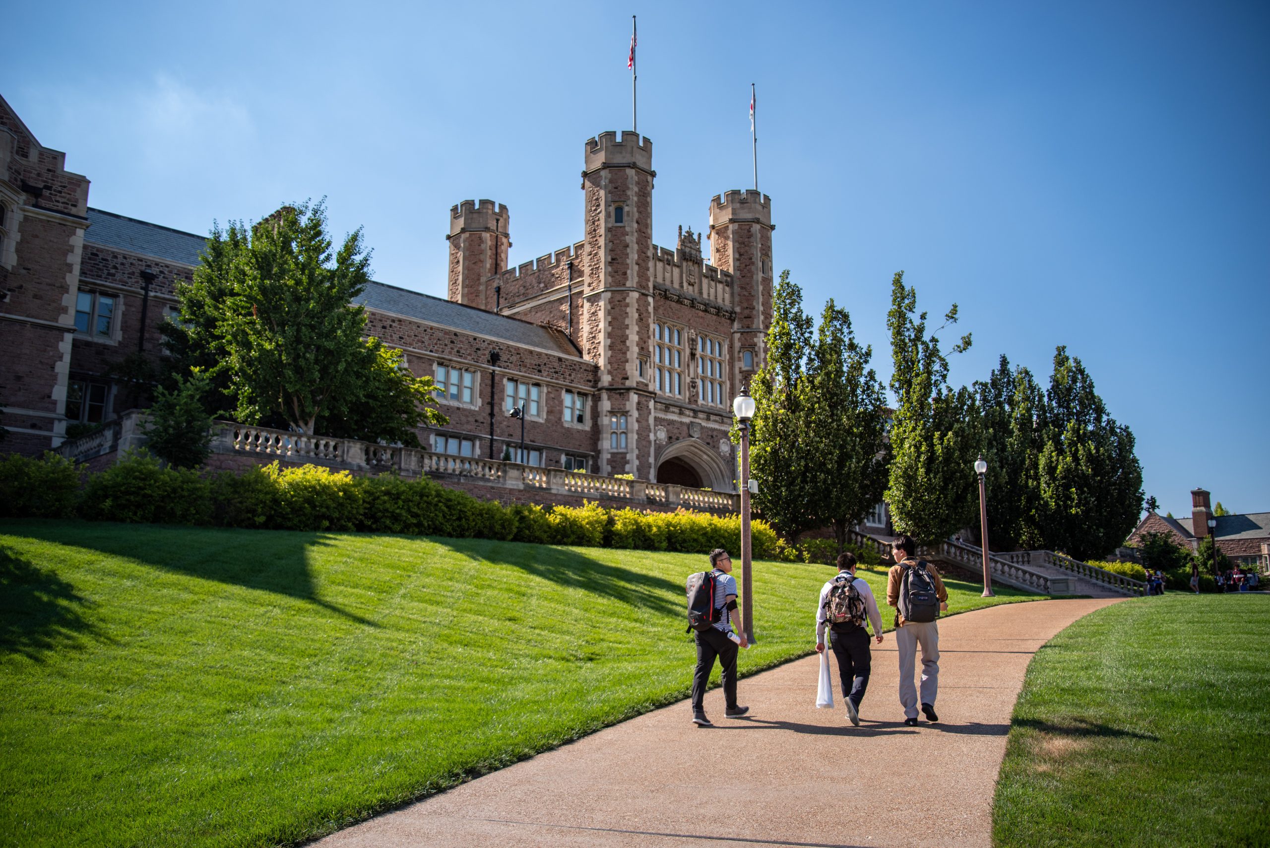 Three people with backpacks on sidewalk in front of the campus administrative building on sunny day moving away.